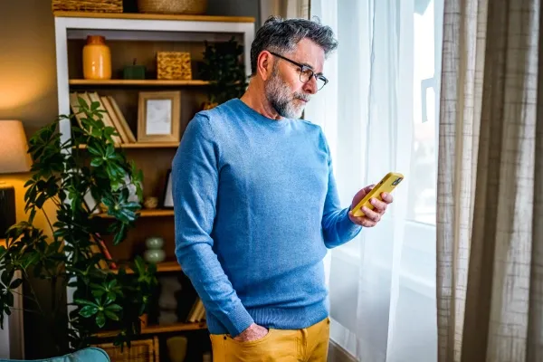 A middle-aged man with a beard and glasses, wearing a light blue sweater and mustard yellow pants, stands by a window and looks intently at a yellow smartphone he holds in his hand.